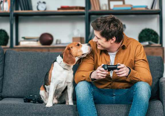 A man is sitting on a grey sofa playing a video game with a dog sitting next to him.