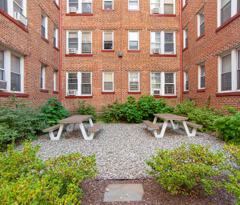 A courtyard with picnic tables and bushes in front of a brick building.
