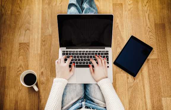 A person is typing on a laptop with a cup of coffee and a tablet beside them.