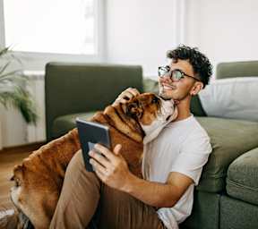 A man is sitting on a green couch with a dog on his lap and using a tablet.