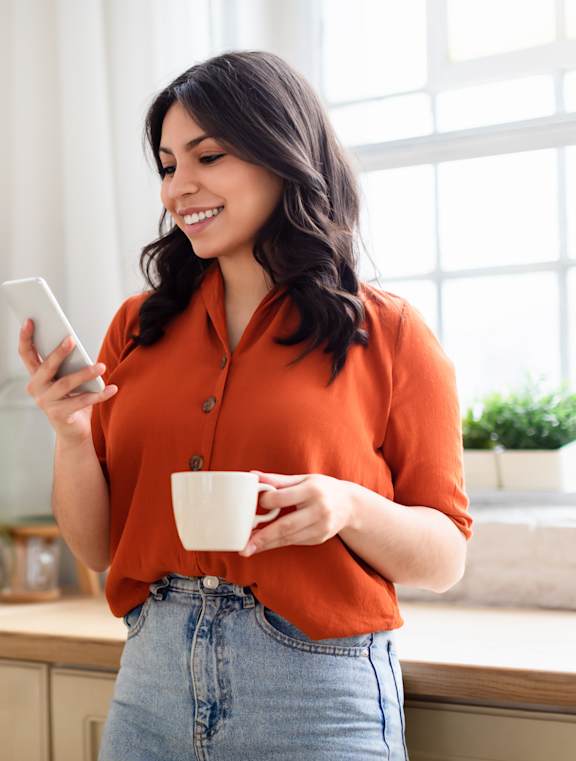 A woman in a red shirt is holding a cup and a phone in a kitchen.