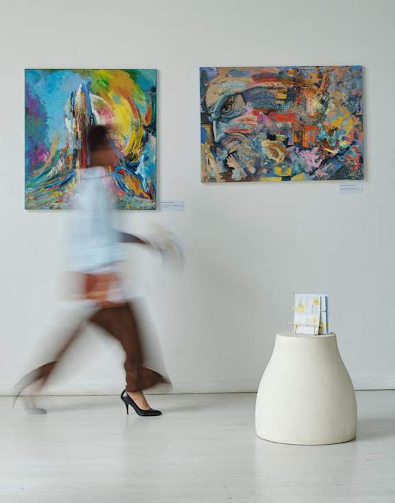 A woman in a white shirt is walking past paintings on a wall.