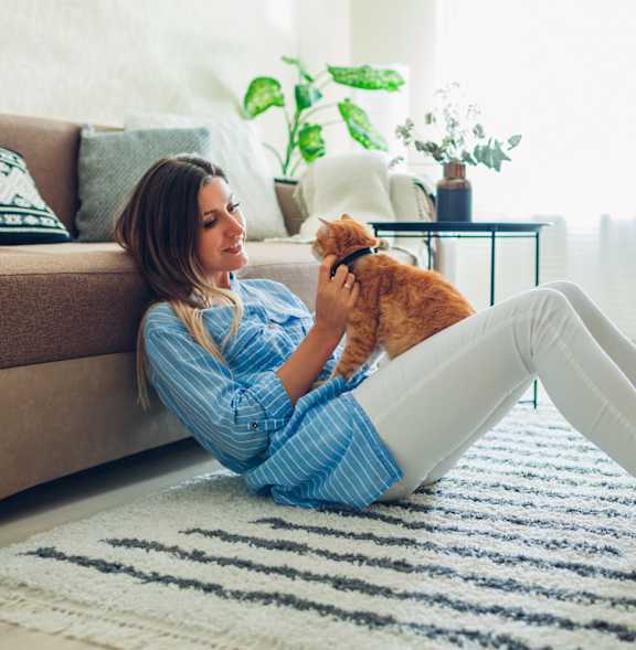A woman in a blue shirt is sitting on a rug with her orange cat.