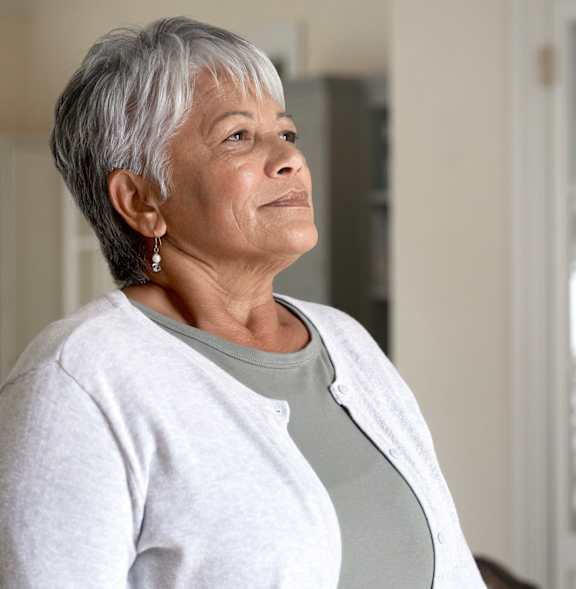 A woman with short grey hair wearing a white cardigan and a grey top stands in front of a white door.