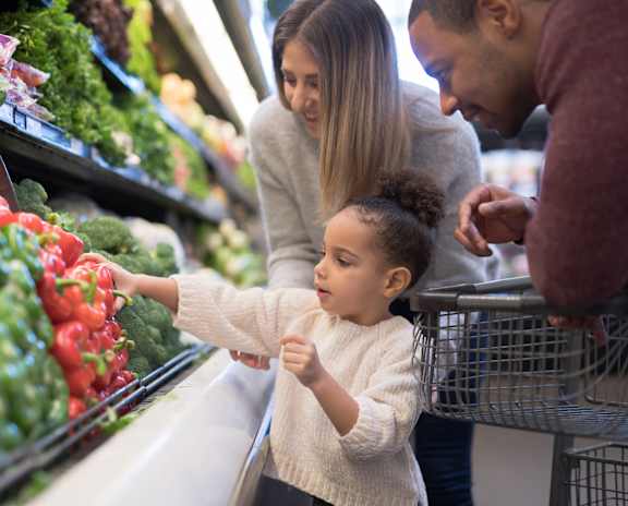 a little girl reaching for a piece of produce at the grocery store with her parents