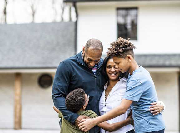A family of four hugging in front of a house.