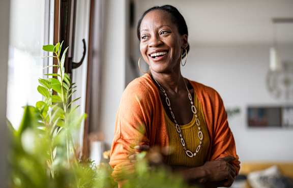 A woman in an orange top is smiling and standing in front of a window with green plants.