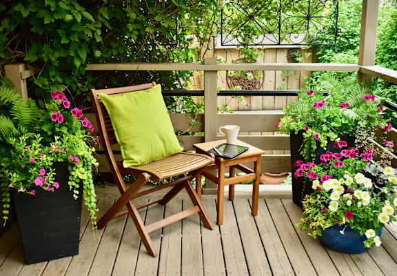 A wooden chair with a green pillow sits on a porch with a table and a book.