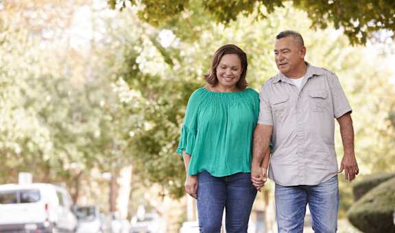 An older couple holding hands and walking down a tree-lined street.