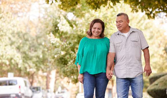 An older couple holding hands and walking down a tree-lined street.