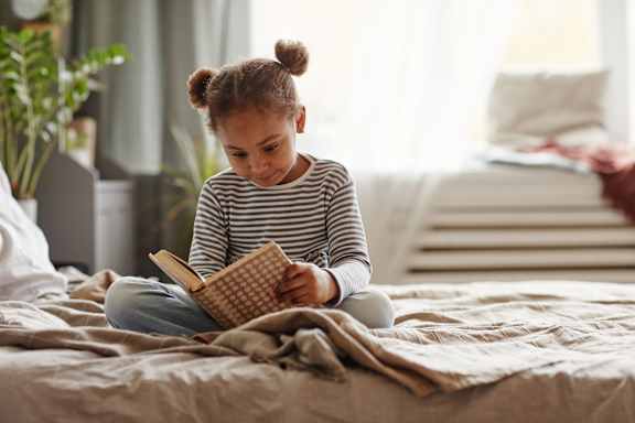 A young girl is sitting on a bed reading a book at The Woodlands Apartments, Milton Keynes, MK5 6GS