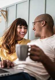 A man and a woman are sitting on a couch, looking at a laptop screen and holding a mug together.
