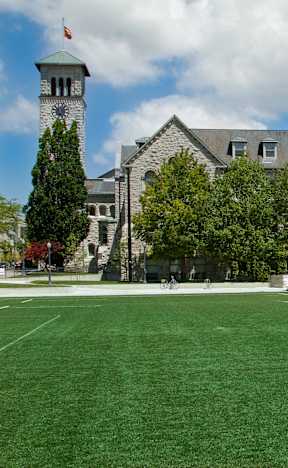 A large building with a tower and a clock is surrounded by trees and has a sports field in front of it.