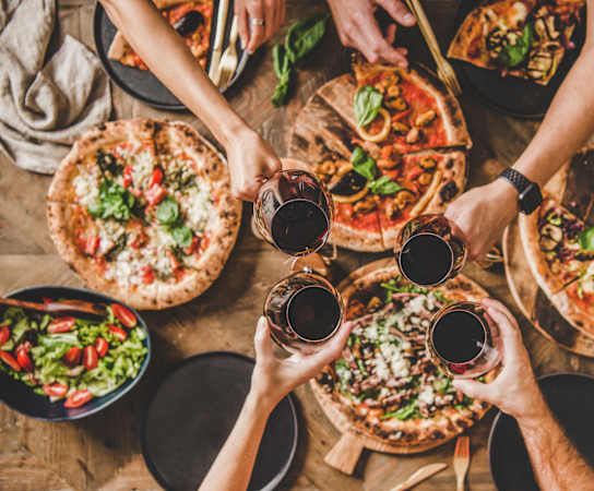 A group of people toasting with glasses of red wine in front of a variety of pizzas.