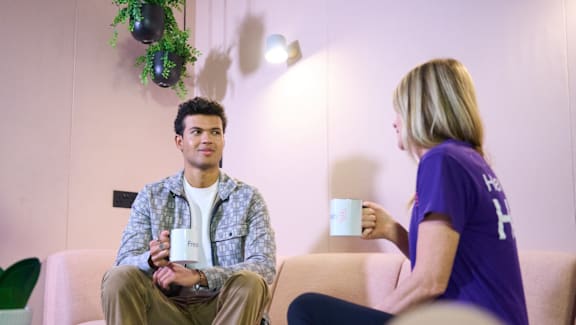 A man and a woman are sitting on a couch, each holding a mug, in a room with a green plant on the side table.