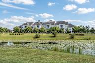 A large white house with a red roof is surrounded by a grassy lawn and a pond with lily pads.