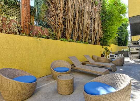 Community Lounge Area with Wicker Seats with Blue Cushions, Wicker Lounge Chairs, Trees, and Yellow Wall at Croft Plaza Apartments, West Hollywood