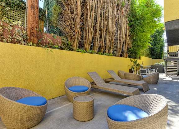 Community Lounge Area with Wicker Seats with Blue Cushions, Wicker Lounge Chairs, Trees, and Yellow Wall at Croft Plaza Apartments, West Hollywood
