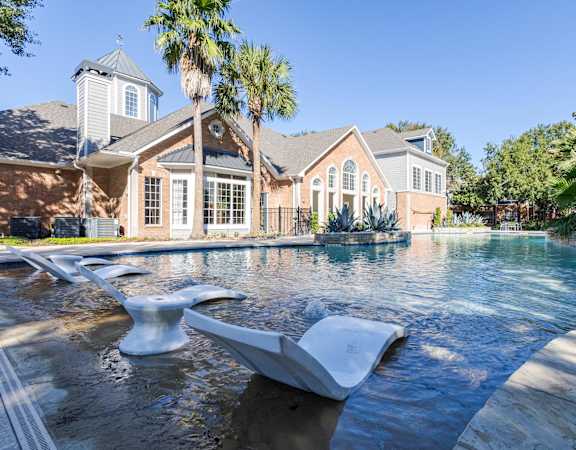 Resort-style pool with in-water lounge chairs, lush palms, and a brick clubhouse with arched windows.
