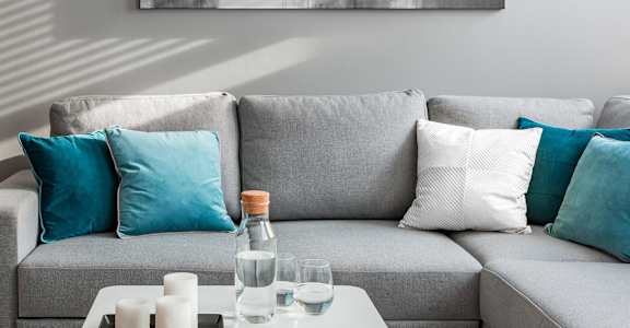 A grey couch with blue and white pillows, a tray with candles, a glass of water, and a bottle of water on a coffee table at Barcelo at East Cobb Apartments, Marietta, GA, 30067