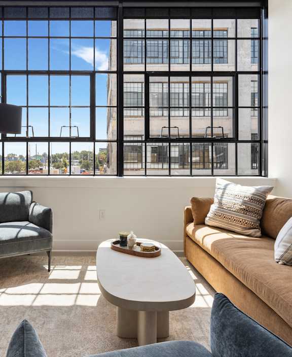 A living room with a brown couch, a grey chair, a wooden cabinet, a white coffee table, and a large window with a view of the outside.