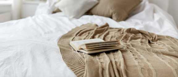 A bed with a white comforter and a brown blanket is in the foreground with a string of lights in the background.