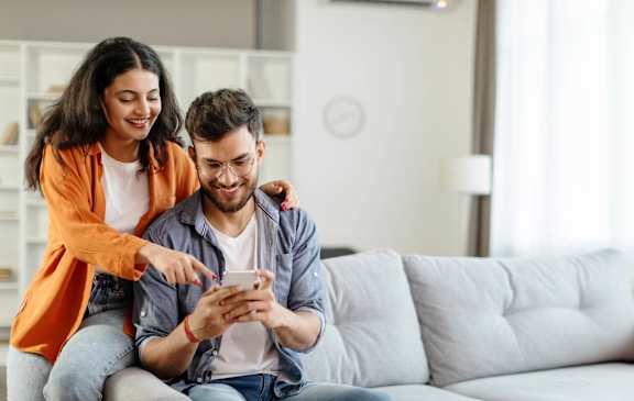 A man and a woman are sitting on a couch, with the woman pointing at the phone the man is holding.