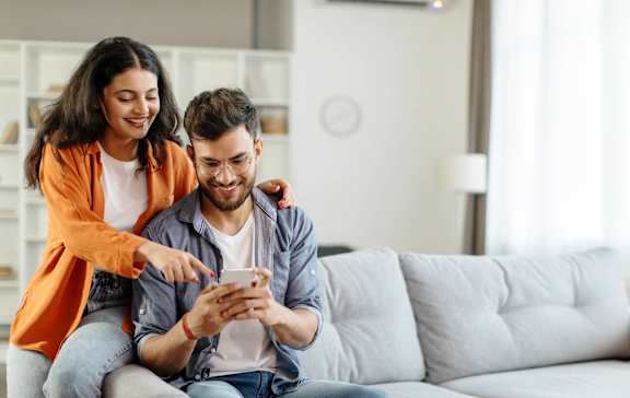 A man and a woman are sitting on a couch, with the woman pointing at the phone the man is holding.