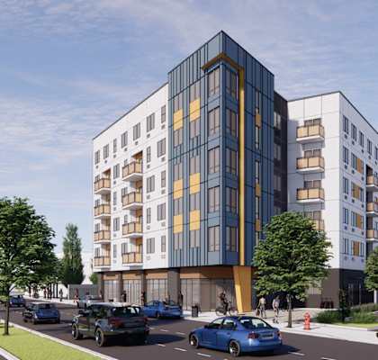 A modern multi-story building with a mix of white and blue panels is surrounded by trees and a street with cars and pedestrians.