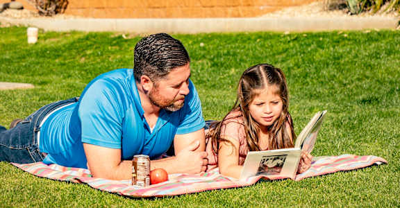 A man and a girl are reading a book on a blanket in the grass.