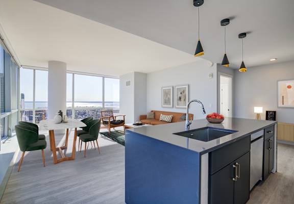 Modern kitchen interior with a central blue island featuring a built-in sink and red apples on top, surrounded by green velvet chairs and set against a bright living space with floor-to-ceiling window