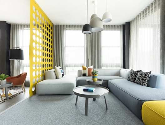 A modern living room with a grey sofa, a yellow accent, and a yellow and black geometric wall panel.