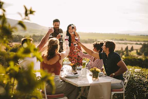 A group of people are gathered around a table in a garden, enjoying a meal and drinks.