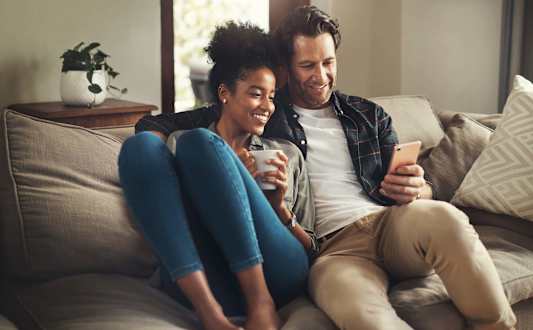 A man and a woman are sitting on a couch, holding hands and smiling at each other at St Helens Place Apartment Homes, St Helens, 97051