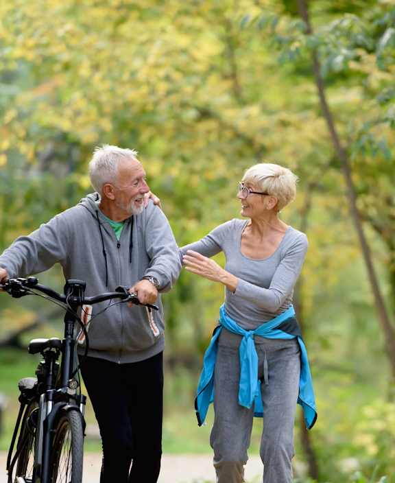 An older couple are walking in the park with a bicycle.