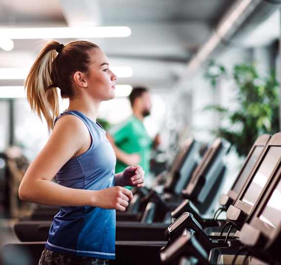 A woman in a blue top and grey shorts is running on a treadmill in a gym.