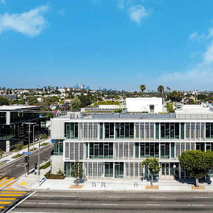 A modern building with a glass facade is situated on a street corner with trees and other buildings in the background.