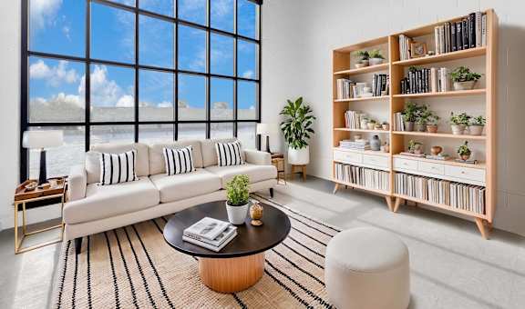 A living room with a large window, a white sofa, a wooden bookshelf, and a coffee table.at Plant 1 Lofts, N. Newton, IA