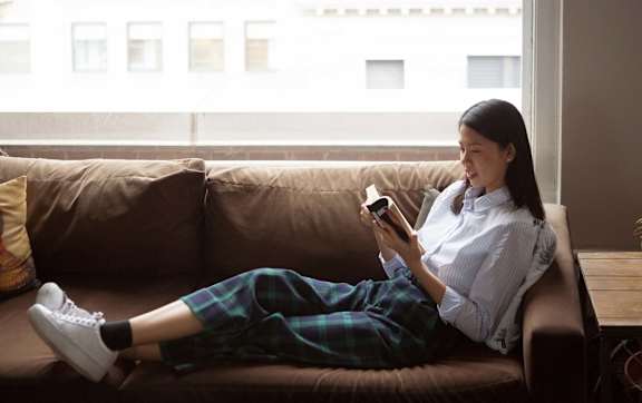 A woman is sitting on a couch with her legs crossed, wearing a white shirt and plaid pants, and holding a phone in her hands.