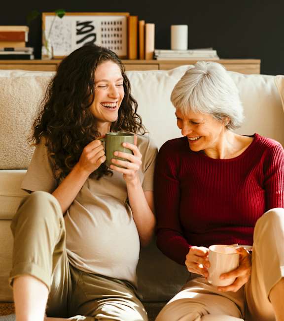 Two women sitting on a couch, one holding a mug and the other holding a cup.
