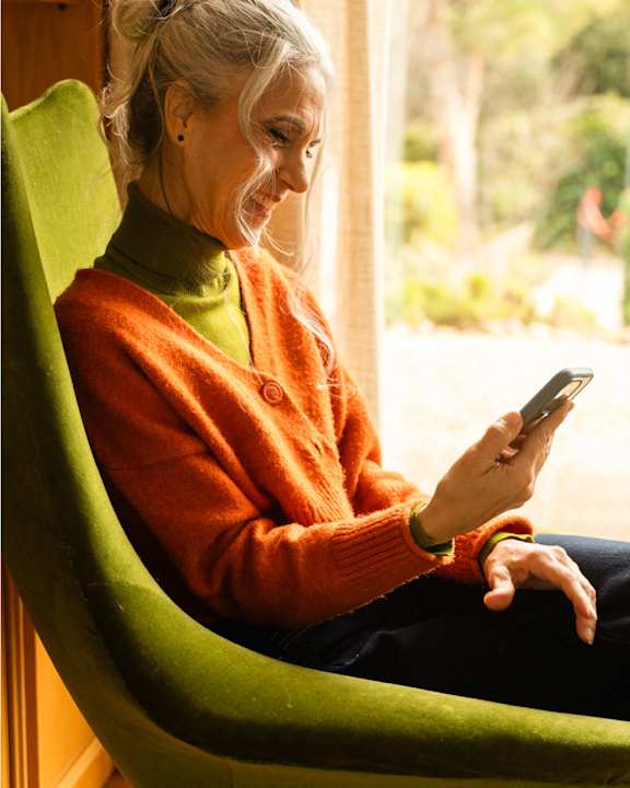 An older woman is sitting in a green chair and looking at her phone.
