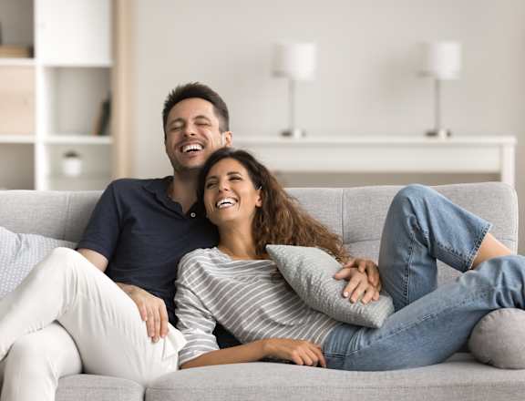 A man and a woman are sitting on a grey sofa, smiling and laughing together.