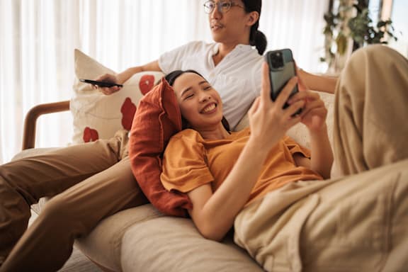 A person is lying on a couch with a pillow, while another person is standing and holding a smartphone.