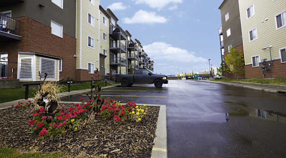 A wet street in front of apartment buildings with a car parked on the side.