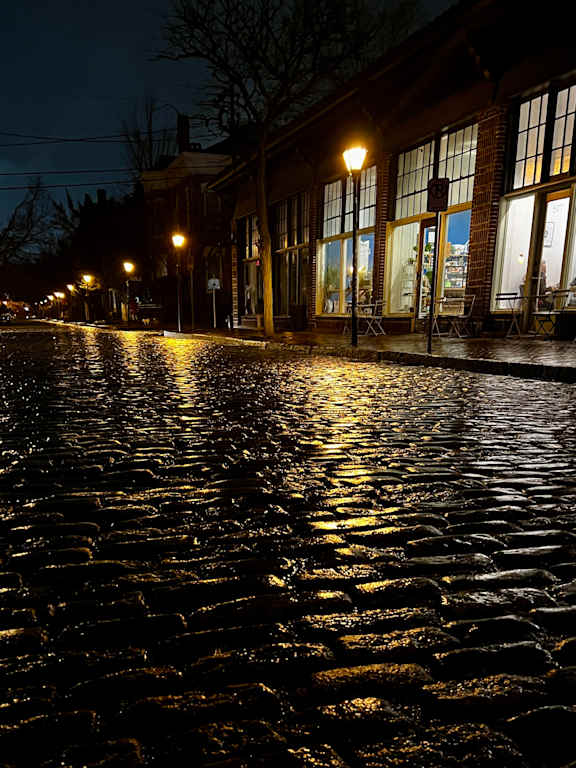 A cobblestone street is lit by street lamps and reflected in the water on the road.