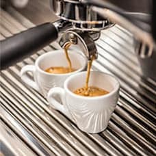Two white cups are being filled with coffee from a silver espresso machine.