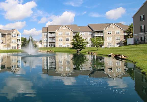 This is a picture of a fountain in a pond with building exteriors at Nantucket Apartments, in Loveland, OH.