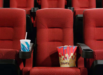 A red cinema seat with a popcorn container and a drink in front of it.
