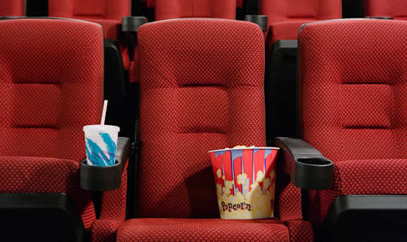 A red cinema seat with a popcorn container and a drink in front of it.