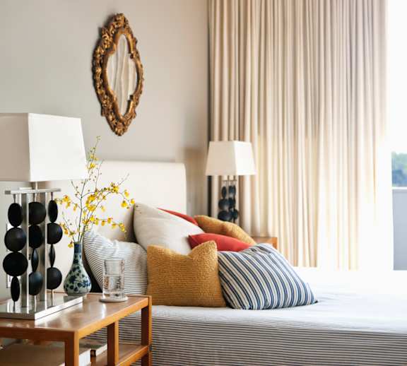 A bedroom with a bed, a nightstand with a lamp, a vase with flowers, and a mirror on the wall.